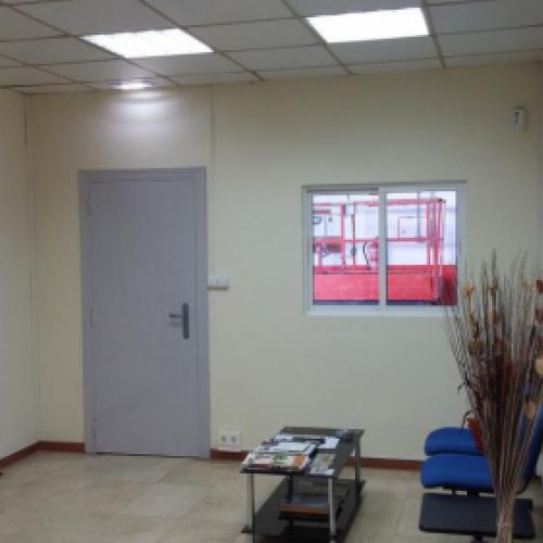 Interior de oficina con planta en primer plano, mesa auxiliar con dos silllas y puerta al fondo