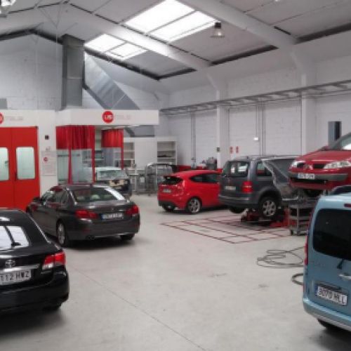 Vista del interior del taller con coches elevados en reparación a la derecha, coches aparcados a la izquierda y puerta de cabina de pintura al fondo