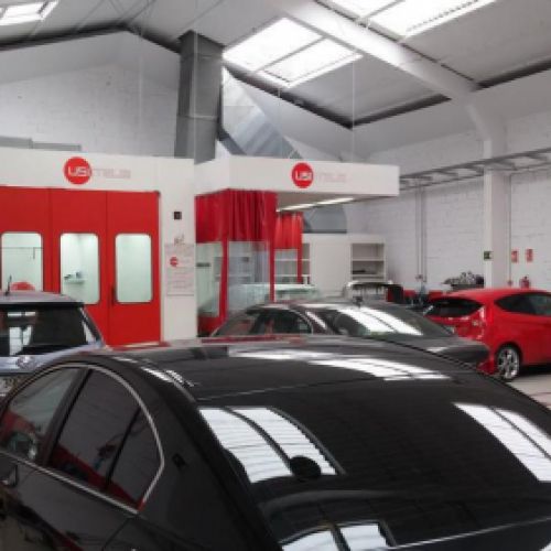Interior del taller con coches aparcados en primer plano y cabina de pintura con puerta roja al fondo