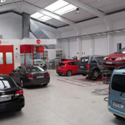 Vista del interior del taller con coches elevados en reparación a la derecha, coches aparcados a la izquierda y puerta de cabina de pintura al fondo