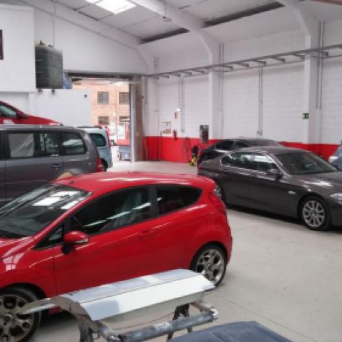 Interior del taller con paredes blancas y zócalo rojo con coches aparcados y puerta de entrada abierta al fondo